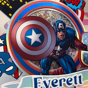 May include: A cake topper featuring Captain America with his shield, a red, white and blue colour scheme, and the name "Everett" on a banner.