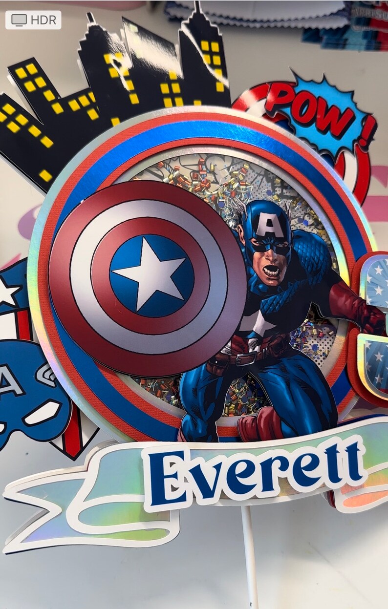 May include: A cake topper featuring Captain America with his shield, a red, white and blue colour scheme, and the name "Everett" on a banner.