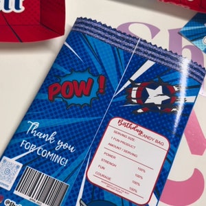 May include: A blue and red party favour bag with a comic book design. The bag features a red "POW!" graphic, a blue and red Captain America shield, and the text "Birthday Candy Bag". The bag also includes a list of "ingredients" such as "power", "strength", "fun", and "courage", all listed as 100%.