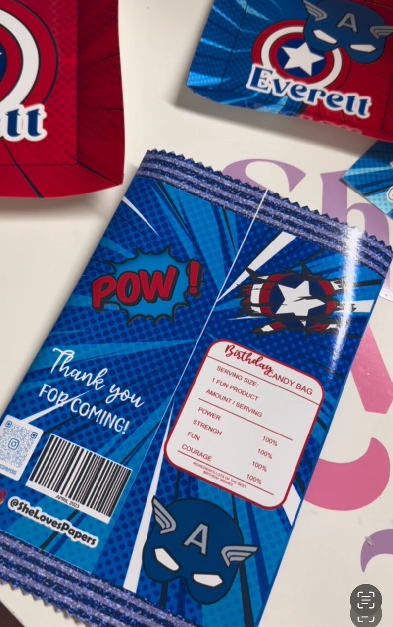 May include: A blue and red party favour bag with a comic book design. The bag features a red "POW!" graphic, a blue and red Captain America shield, and the text "Birthday Candy Bag". The bag also includes a list of "ingredients" such as "power", "strength", "fun", and "courage", all listed as 100%.