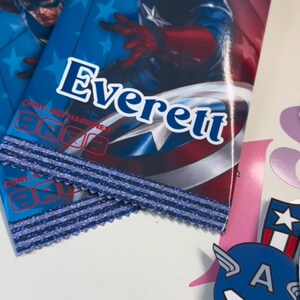 May include: Two blue and red Captain America themed party favour bags with the name Everett printed on the front. The bags have a blue and silver glitter trim.