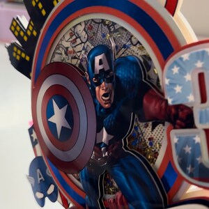 May include: A red, white and blue cake topper featuring Captain America with his shield, a cityscape, and the name "Everett".