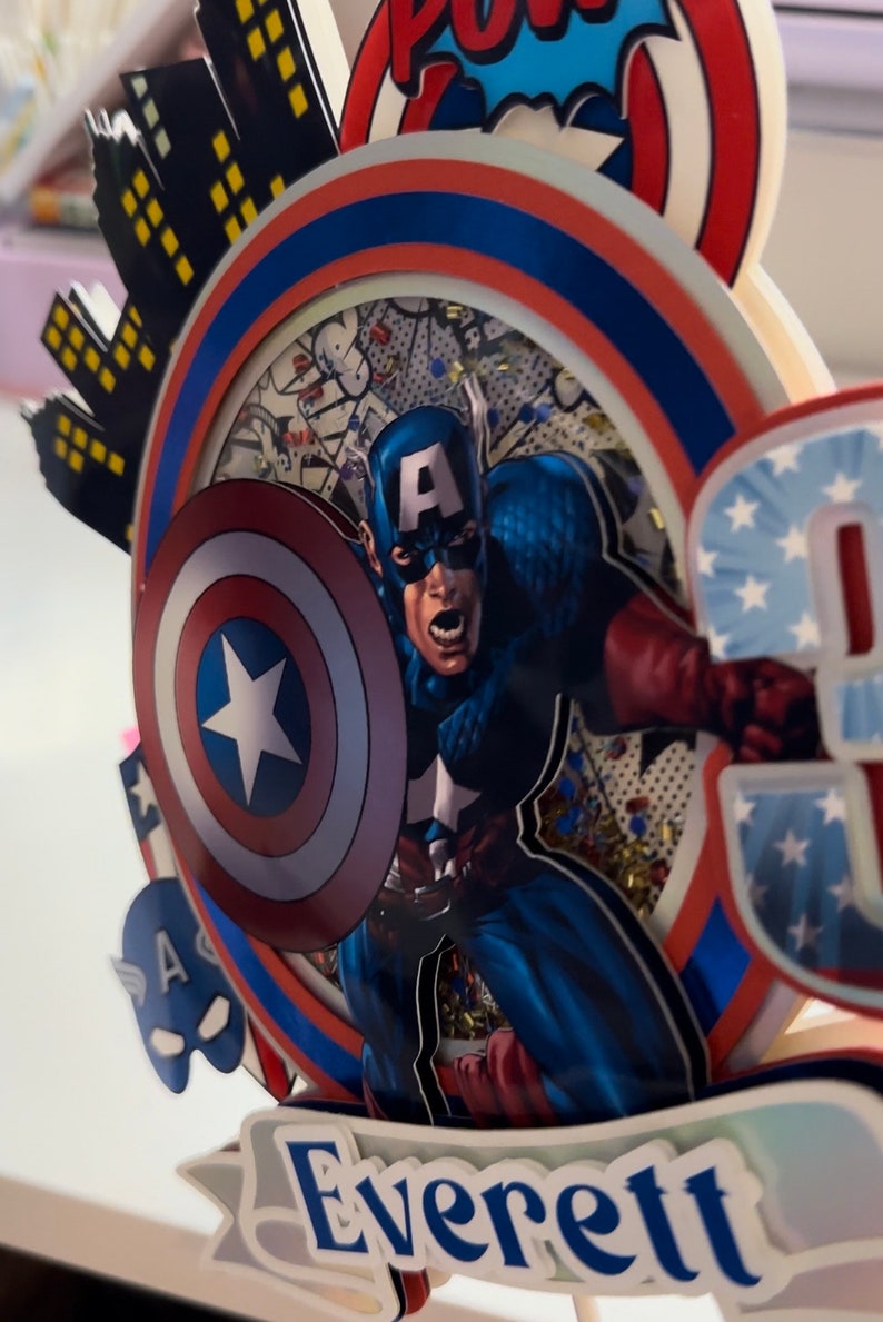 May include: A red, white and blue cake topper featuring Captain America with his shield, a cityscape, and the name "Everett".