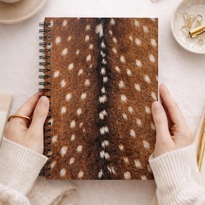 Può includere: Quaderno a spirale con un motivo maculato marrone e bianco, simile alla pelliccia di cervo. Il quaderno ha una rilegatura a spirale nera ed è tenuto da una persona che indossa un maglione color crema. Lo sfondo include una tazza di caffè e articoli di cancelleria.