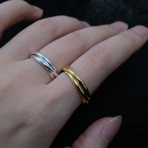 Peut inclure: Deux bagues entrelacées, une argentée et une dorée, sont présentées. Les bagues ont une finition lisse et polie et un design simple et élégant. La bague dorée est sur un doigt, tandis que la bague argentée est sur un autre doigt.