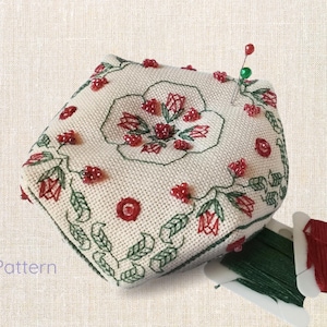 Peut inclure: Coussin à épingles crème avec un motif floral brodé en rouge et vert. Le coussin présente un motif floral central et des perles rouges. Deux épingles sont insérées sur le dessus. Le texte "PDF Pattern" est visible.