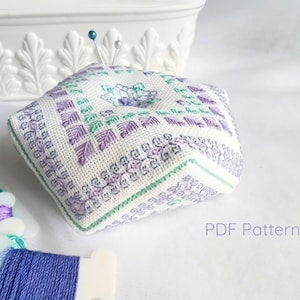 May include: Square white pin cushion with a floral and geometric pattern in shades of purple and green. Two pins are inserted into the cushion. Embroidery floss in shades of purple, blue, and green are in the foreground. The text "PDF Pattern" is visible.