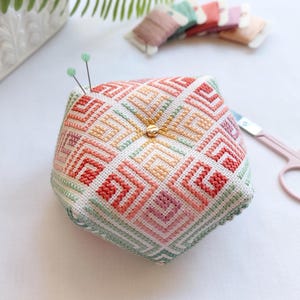 Op de afbeelding: Een vijfhoekig speldenkussen met een geometrisch kruissteekpatroon in tinten koraal, perzik, roze en mintgroen. Het midden is versierd met een gouden kraal en twee spelden met lichtgroene kopjes zijn in de bovenkant van het kussen gestoken.