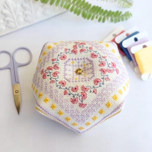 Peut inclure: Un pelote à épingles pentagonale avec un dessus en tissu crème orné d'un motif floral au point de croix rose, violet et jaune, centré par une petite épingle dorée en forme d'étoile. À côté se trouvent des ciseaux violets et des bobines de fil.