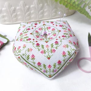 Peut inclure: Coussin à épingles blanc en forme de pentagone, avec un motif floral brodé en rose et vert. Deux épingles sont insérées. Du fil à broder rose et des ciseaux roses sont également visibles.