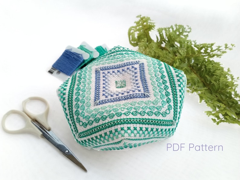 Hand-embroidered biscornu pincushion in shades of green, turquoise, and blue, featuring a geometric design with a decorative central square. Surrounded by embroidery floss bobbins, small scissors, and a sprig of fern.  Text indicating that is a a digital product.