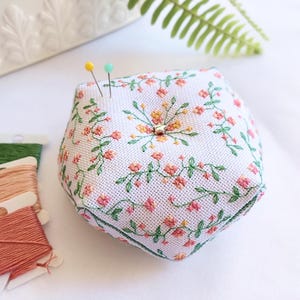 Puede incluir: Un acerico pentagonal de tela blanca con enredaderas verdes bordadas y pequeñas flores rosas y amarillas. Una pequeña cuenta plateada en forma de estrella está centrada en la parte superior, con dos alfileres insertados. Tres carretes de hilo están cerca.