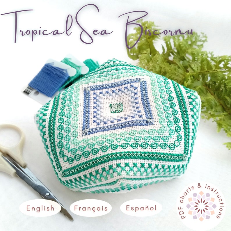 Hand-embroidered biscornu pincushion in shades of green, turquoise, and blue, featuring a geometric design with a decorative central square. Surrounded by embroidery floss bobbins, small scissors, and a sprig of fern. In the top left corner, there's a floral logo with the text 'PDF charts & instructions,' and at the bottom right, the available languages are listed: English, Français, Español.