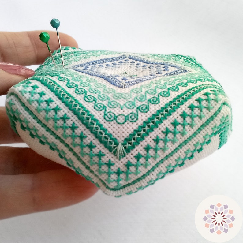 Lateral view showing corners of an octagonal embroidered pin cushion with sea wave motifs in varying shades of green and blue, complemented by pearly sea foam-colored thread ornaments. Hold in hand for size reference.