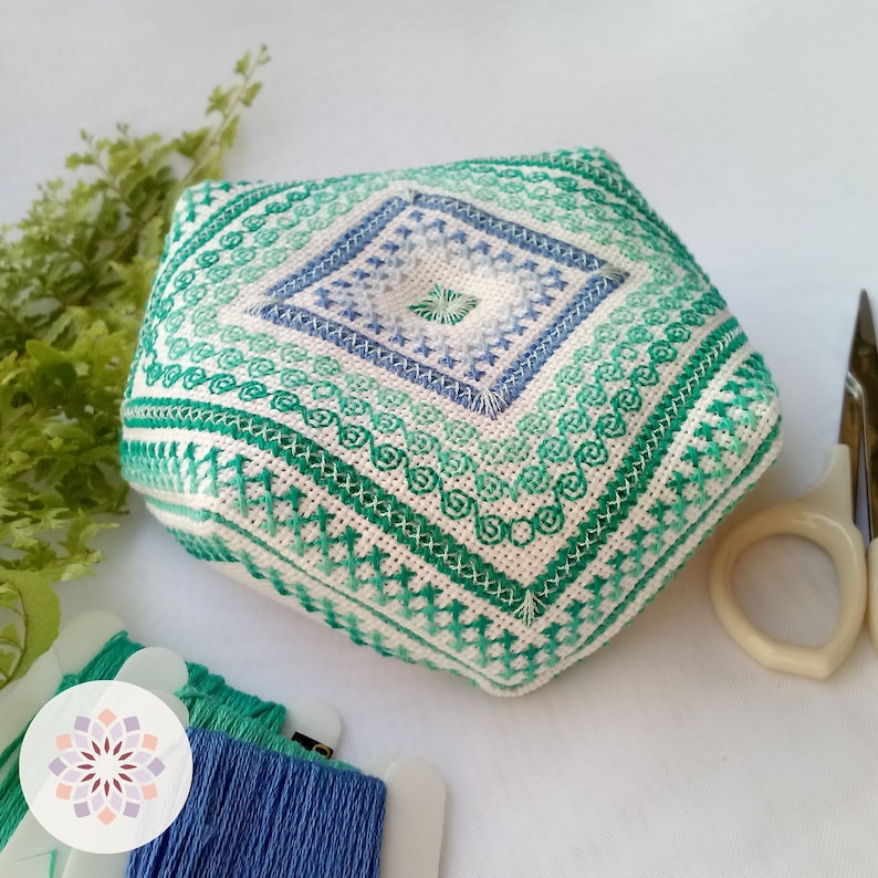 Detailed view of octagonal pin cushion, adorned with vibrant sea wave patterns in greens and blues. Accented with luminescent sea foam-colored thread ornaments, reminiscent of ocean waves. Alternating corners details.