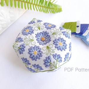 Op de afbeelding: Een zeshoekig speldenkussen met een wit bloemenpatroon met blauwe bloemen en groene accenten. Spoelen met groen en blauw borduurgaren liggen ernaast. De tekst "PDF Pattern" is zichtbaar.