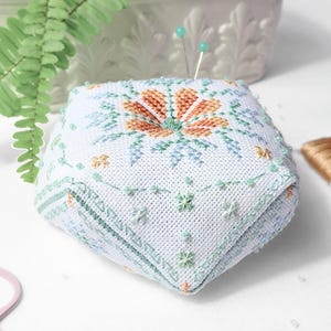 May include: A white, square pincushion with a floral design in orange and green cross-stitch. The pincushion has a geometric shape and is decorated with small, embroidered flowers and beads. Two pins are inserted into the top. Sewing supplies are in the background.