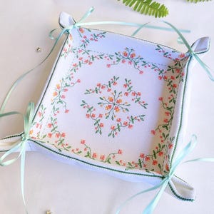 Peut inclure: Un vide-poche carré en tissu avec une base blanche ornée d'un motif brodé de petites fleurs roses et de vignes vertes. Les coins sont froncés et maintenus par des rubans de satin vert clair noués en nœuds à chaque extrémité.
