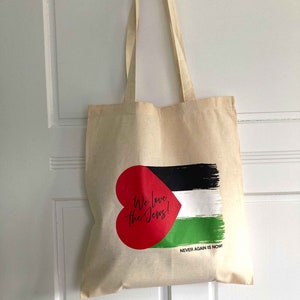 Könnte beinhalten: Eine beige Tragetasche mit einem roten Herzen und dem Text "We love the Jews!" darauf gedruckt. Das Herz ist teilweise von einer schwarz-weiß-grünen Flagge bedeckt, unter der der Text "Never Again is Now!" steht.