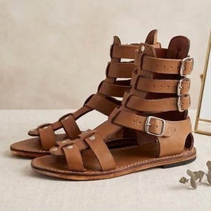 May include: A pair of tan leather gladiator sandals featuring multiple horizontal straps across the foot and ankle. Each sandal includes four adjustable silver-toned metal buckles on the outer side, a flat sole, and visible stitching along the edges.