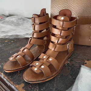 May include: A pair of tan leather gladiator sandals featuring a high-cut design with multiple horizontal straps, silver-toned metal buckles, and small silver rivets securing the straps to the base. The sandals have a flat sole and open toe.