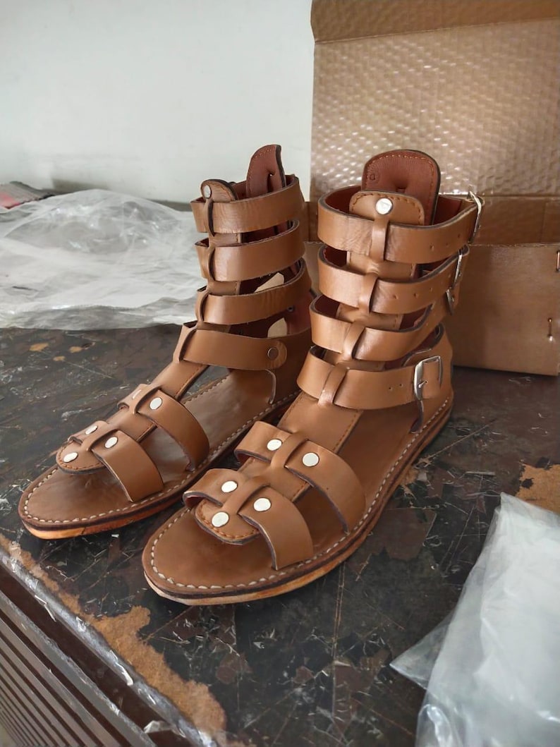 May include: A pair of tan leather gladiator sandals featuring a high-cut design with multiple horizontal straps, silver-toned metal buckles, and small silver rivets securing the straps to the base. The sandals have a flat sole and open toe.