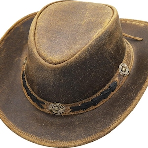Può includere: Cappello da cowboy in pelle marrone invecchiata con corona arrotondata e tesa larga. Il cappello presenta una fascia decorativa con cuciture nere e due conchos metallici. Un cinturino in pelle è attaccato alla tesa.