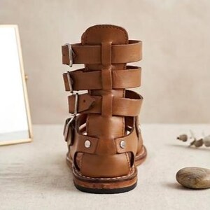 May include: A tan leather gladiator sandal featuring a high-cut back, four horizontal straps with silver-toned metal buckles, and a flat sole. The design includes a vertical center strap connecting the horizontal bands and visible silver rivets.
