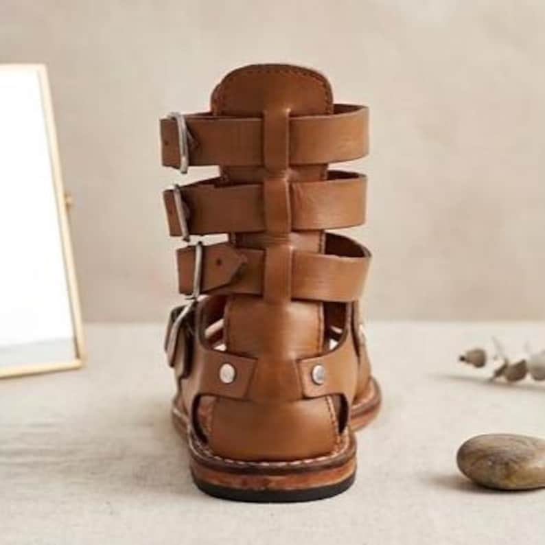 May include: A tan leather gladiator sandal featuring a high-cut back, four horizontal straps with silver-toned metal buckles, and a flat sole. The design includes a vertical center strap connecting the horizontal bands and visible silver rivets.