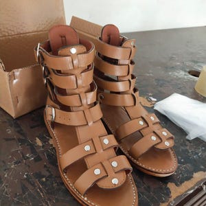 May include: A pair of tan leather gladiator sandals featuring multiple horizontal straps, silver-toned metal buckles, and small silver rivets. The sandals have a high-cut ankle design with a flat sole and visible white stitching along the edges.
