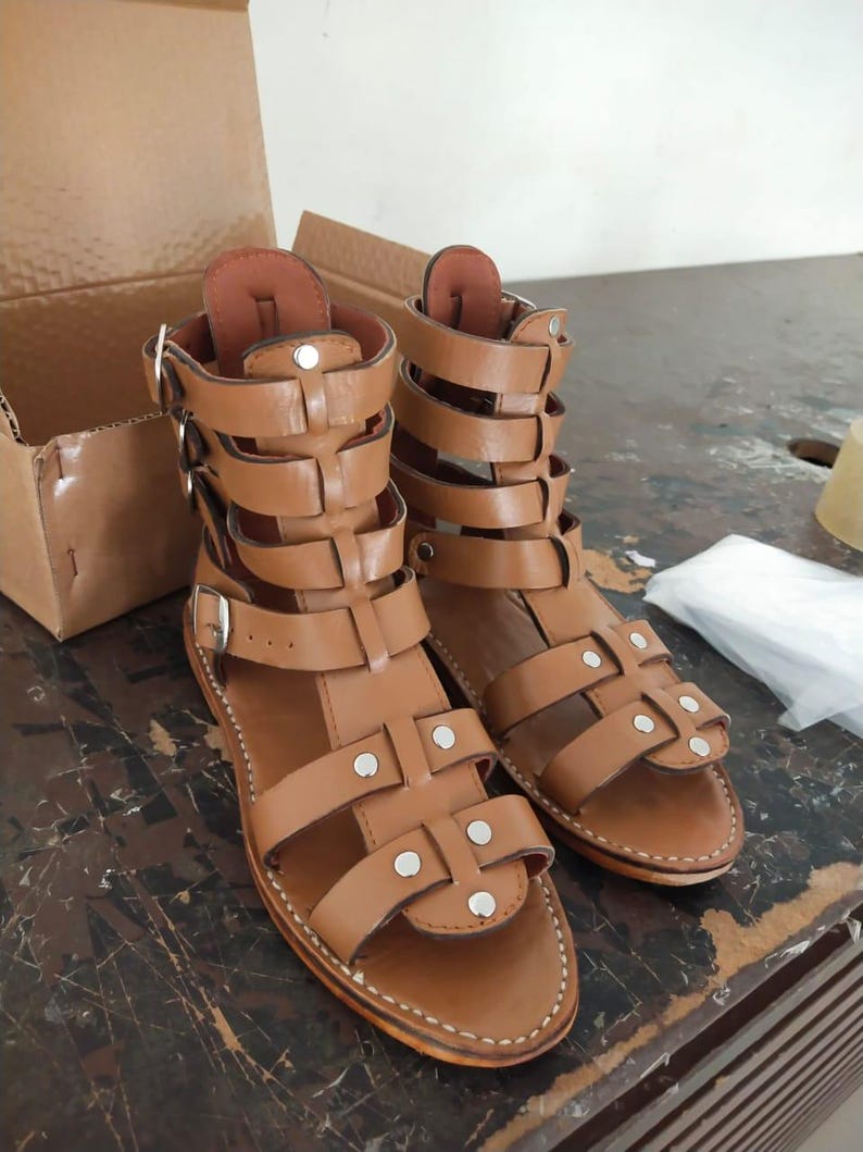 May include: A pair of tan leather gladiator sandals featuring multiple horizontal straps, silver-toned metal buckles, and small silver rivets. The sandals have a high-cut ankle design with a flat sole and visible white stitching along the edges.