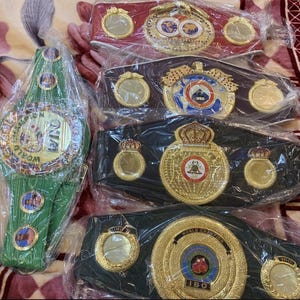 Puede incluir: Una variedad de cinturones de campeonato de boxeo en rojo, verde y negro. Cada cinturón presenta una hebilla dorada grande y ornamentada con diseños y texto intrincados, envueltos en plástico transparente.