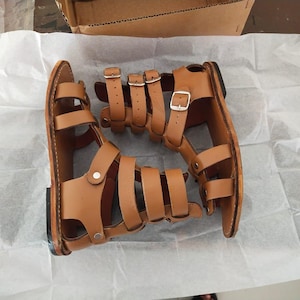 May include: A pair of tan leather gladiator sandals featuring multiple horizontal straps across the foot and ankle, secured with silver-toned metal buckles and rivets. The sandals have a flat sole and an open-toe design with a high-cut ankle strap.