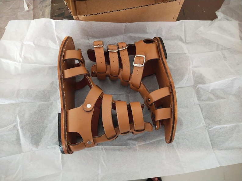 May include: A pair of tan leather gladiator sandals featuring multiple horizontal straps across the foot and ankle, secured with silver-toned metal buckles and rivets. The sandals have a flat sole and an open-toe design with a high-cut ankle strap.