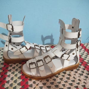 Peut inclure: Une paire de sandales blanches de style gladiateur avec de multiples lanières et des boucles en métal couleur bronze. Ces sandales montantes possèdent une fermeture éclair à l'arrière et une semelle plate en bois clair. Les lanières entourent le pied.