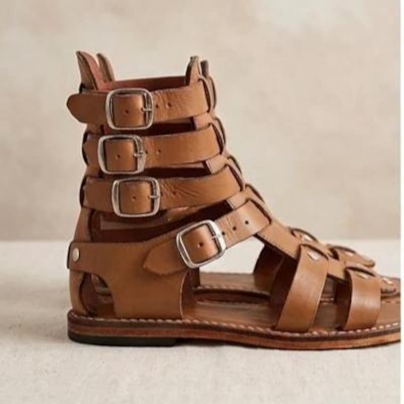 May include: A tan leather gladiator sandal featuring a high-cut ankle design with four horizontal straps, each secured by a silver-toned metal buckle. The sandal has a flat sole with visible stitching along the edge and an open-toe construction.