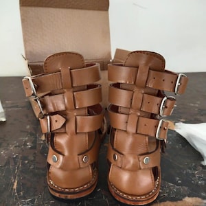 May include: A pair of tan leather gladiator-style sandals featuring three horizontal straps with silver-toned metal buckles on each side. The sandals have a high-cut design with a reinforced heel and silver rivets at the base, set against a cardboard box.