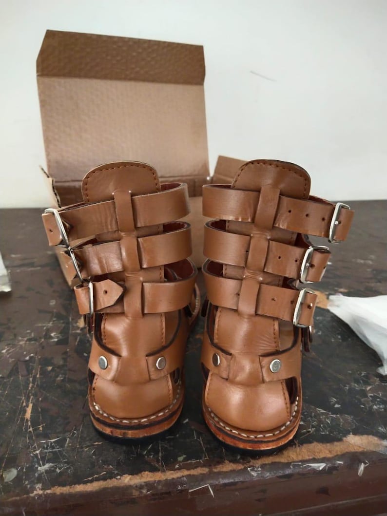 May include: A pair of tan leather gladiator-style sandals featuring three horizontal straps with silver-toned metal buckles on each side. The sandals have a high-cut design with a reinforced heel and silver rivets at the base, set against a cardboard box.