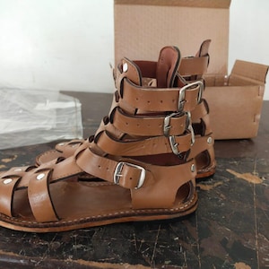 May include: A pair of tan leather gladiator sandals featuring multiple straps that wrap around the ankle and foot, secured with silver-toned metal buckles and rivets. The sandals have a flat sole and an open-toe design with a high-cut ankle structure.