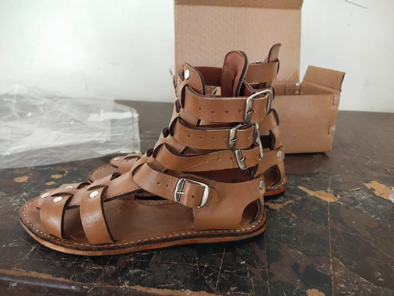 May include: A pair of tan leather gladiator sandals featuring multiple straps that wrap around the ankle and foot, secured with silver-toned metal buckles and rivets. The sandals have a flat sole and an open-toe design with a high-cut ankle structure.
