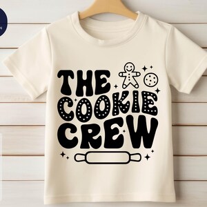 May include: White t-shirt with black text that reads "The Cookie Crew" and a gingerbread man graphic. There is also a rolling pin graphic and a cookie graphic.