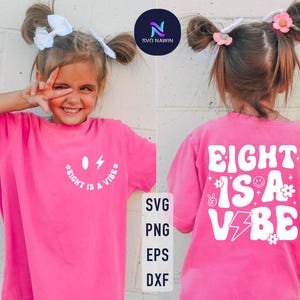 Puede incluir: Una camiseta rosa brillante con el texto blanco "EIGHT IS A VIBE". La parte delantera tiene un diseño de cara sonriente y rayo. La parte trasera de la camiseta también tiene el texto "EIGHT IS A VIBE" con gráficos adicionales.
