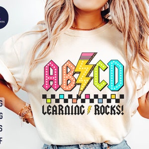 May include: A white t-shirt with a colorful graphic design featuring the letters "ABCD" in a retro style with a lightning bolt and a pencil. The text "Learning Rocks!" is below the letters.