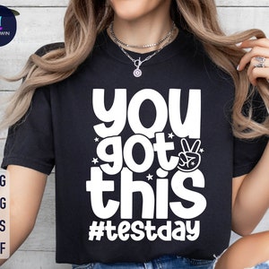 May include: Black t-shirt with white text that reads "You got this #testday" and a peace sign graphic.