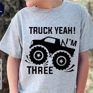 Può includere: Maglietta grigia con una grafica di un camion mostro nero e il testo "Truck Yeah! I'm Three".