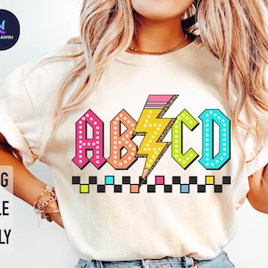 Könnte beinhalten: Ein weißes T-Shirt mit einem bunten Grafikdesign, das die Buchstaben "ABCD" im Retro-Stil mit Punkten und einem Blitz zeigt. Die Buchstaben sind schwarz umrandet und haben ein kariertes Muster darunter.