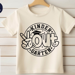 Puede incluir: Camiseta blanca con un gráfico circular negro que dice "Kinder Out Garten" con un gorro de graduación y un signo de paz.