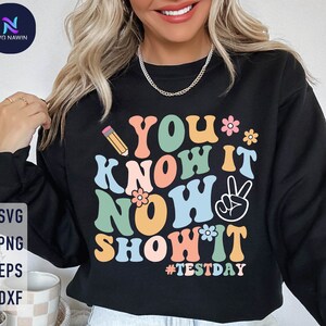 May include: Black sweatshirt with the text "YOU KNOW IT NOW SHOW IT" in colorful retro font, a pencil, flowers, and a peace sign. The hashtag #TESTDAY is also included. The design is perfect for test day.