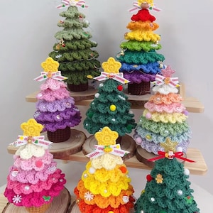 Könnte beinhalten: Eine Sammlung handgefertigter gehäkelter Weihnachtsbäume in verschiedenen Farben, darunter Grün, Rosa und Regenbogenfarben. Jeder Baum ist mit einem gelben Sternaufsatz, dekorativen Schleifen und kleinen Verzierungen geschmückt. Die Bäume werden auf Holzplattformen ausgestellt.