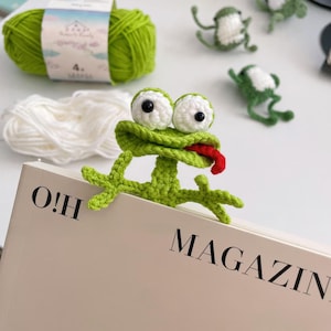 Puede incluir: Marcapáginas de rana verde hecho a mano con ganchillo, con ojos blancos y lengua roja, colocado en un libro. El libro tiene el texto "O!H MAGAZINE". Se ven ranas pequeñas de ganchillo adicionales en el fondo, junto con hilo verde.